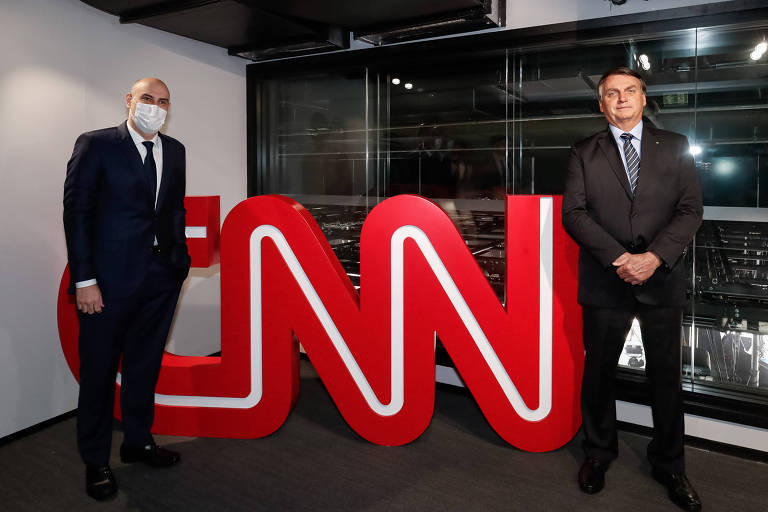 Dois homens brancos usam roupa formal preta e aparecem entre letreiro grande, onde está escrito "CNN"