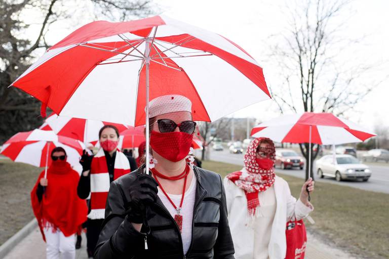 Mulheres caminham em grupo na calçada com guarda-chuvas vermelhos e brancos abertos