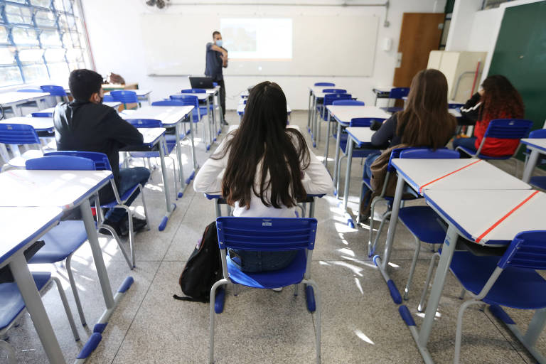 Em uma sala de aula, estudantes de costas estão sentados distantes um dos outros.
