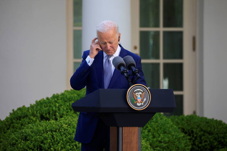 Biden, de terno azul, em frente a púlpito, coloca o dedo na orelha para escutar melhor uma traduçãp
