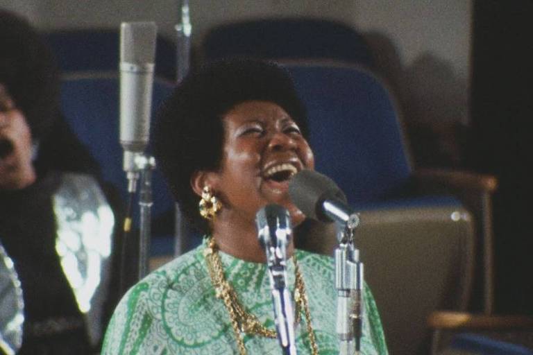 Mulher negra usa roupa verde e canta com microfone