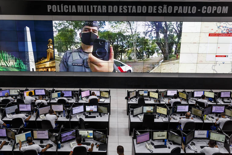 Central de monitoramento mostra sala de computadores e um telão com a imagem de um policial mostrando a câmera 