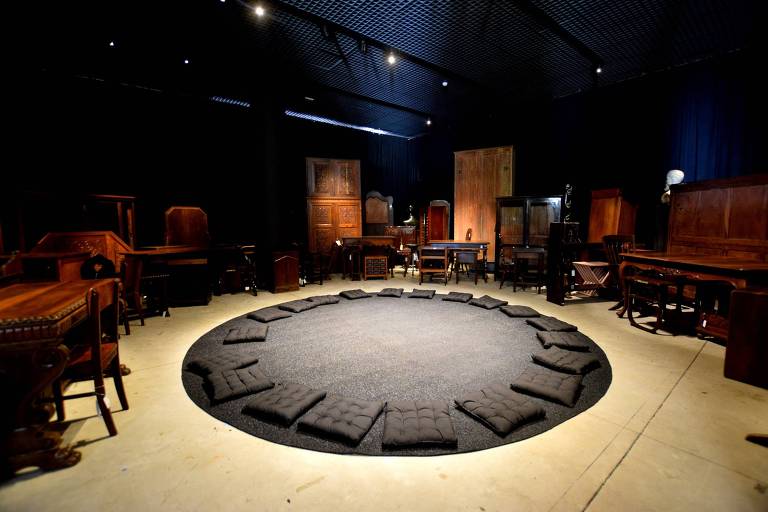 instalação circular tem móveis de madeira antigos espalhados por uma sala e, ao centro do recinto, um tapete preto com almofadas em círculo