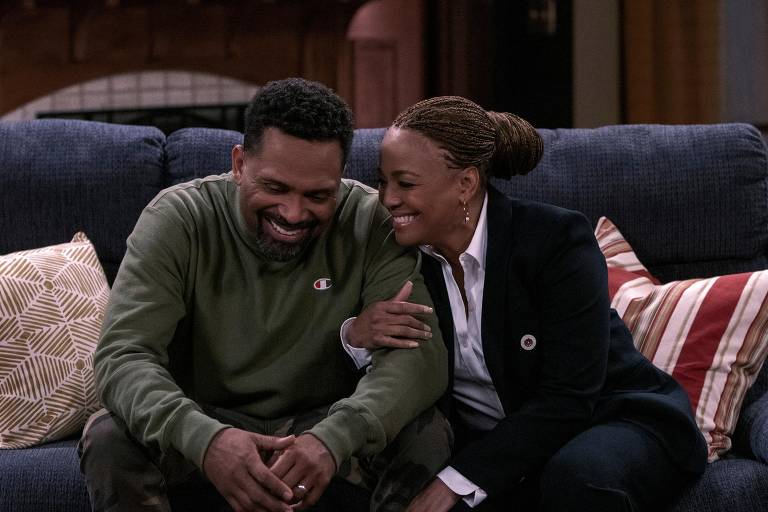 Um casal negro está sentado num sofá; os dois riem, e ela segura o braço esquerdo dele