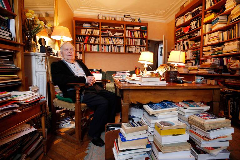 Homem branco idoso sentado em frente à pilha de livros e estante 