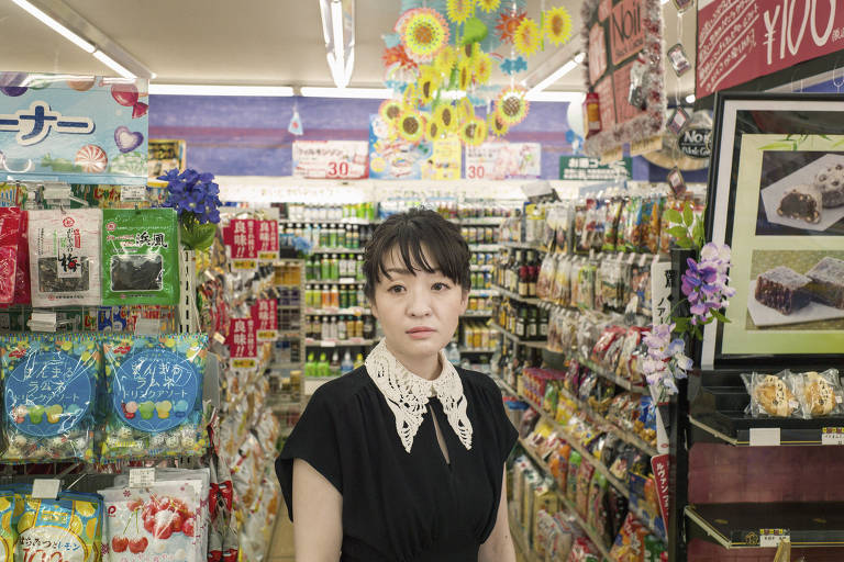 A escritora Sayaka Murata, fotografada em uma konbini em Tóquio; ela usa um vestido preto com uma gola de renda branca; olha para a câmera de frente, com olhar meio perdido, em meio a prateleiras multicoloridas com produtos variados, como lanches, à venda na loja de conveniência