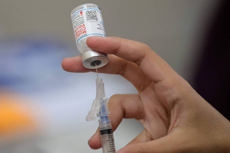 Uma pessoa insere uma seringa com agulha em uma ampola da vacina da Moderna contra Covid-19 ao mesmo tempo que puxa o êmbolo para preencher o líquido em primeiro plano; ao fundo, uma parede cinza.