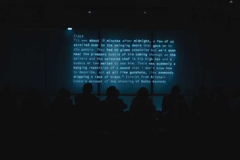 Em sala escura, público olha para tela, que mostra depoimentos por escrito