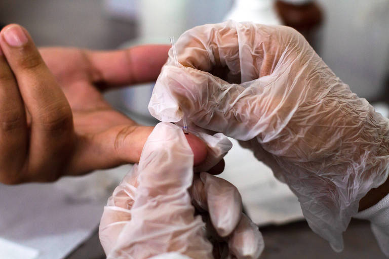 Imagem mostra as mãos de profissional de saúde, com luvas, lancetando o dedo de um paciente