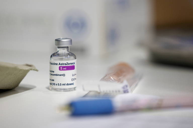 Frasco com doses da vacina da AstraZeneca está em cima de uma mesa branca, perto de um saquinho plástico com inscrições e um objeto azul que aparece desfocado