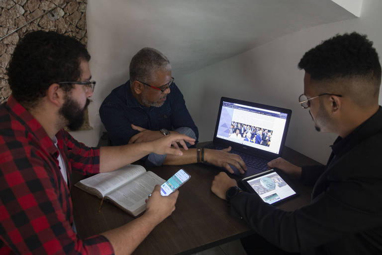 Três homens sentados à mesa, onde há um computador e uma Bíblia. Um deles usa o celular e outro um tablet que mostram a página do observatório evangélico