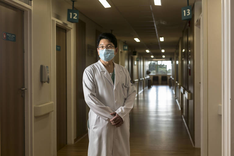 De jaleco branco e máscara, homem de cabelos pretos posa em corredor de hospital