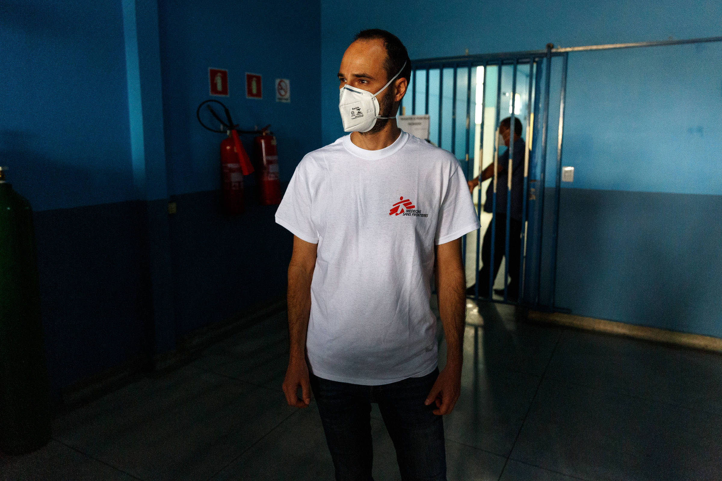 Homem de camiseta branca e máscara em frente a parede azul