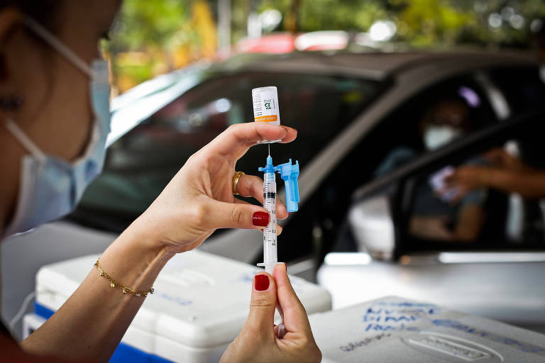 Em primeiro plano, enfermeira de máscara segura seringa e frasco de vacina; ao fundo, carro em que pessoa é vacinada