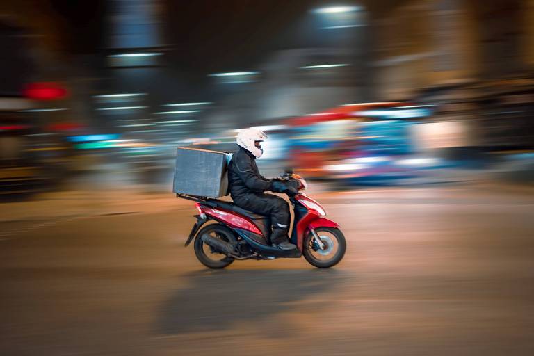 Homem agasalhado, de moto vermelha, com capacete branco, faz entregas numa cidade à noite