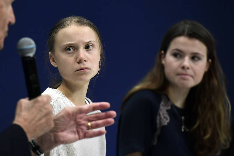 Duas garotas olham para pessoa que fala ao microfone do lado esquerdo da imagem (apenas a mão e o microfone dele aparecem na foto)