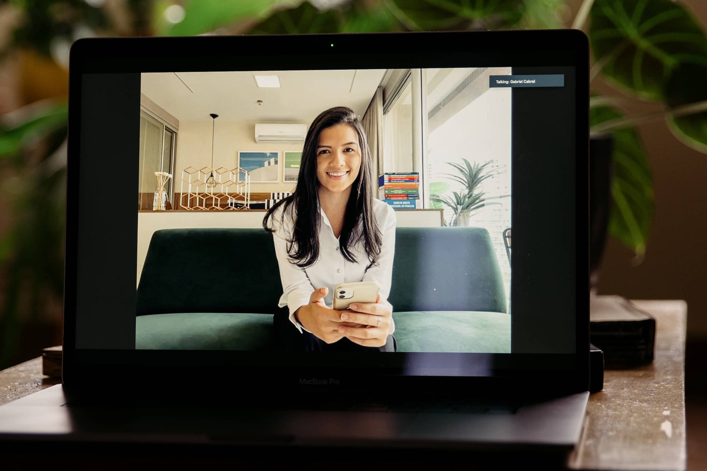Tela de computador onde se vê mulher branca sorrindo e com braço esticado para segurar o celular com que se filma. Ao fundo ambiente de escritório