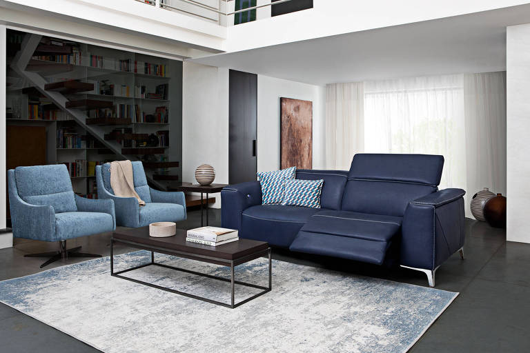 Sala de estar, com um sofá azul escuro para duas pessoas, e dois sofás individuais azul claro. Há um tapete cinza e uma mesa de centro.