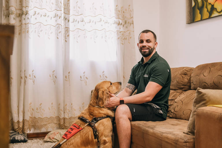 Homem de camiseta e bermuda sentado no sofá, acariciando uma golden retriever com equipamento de cão guia