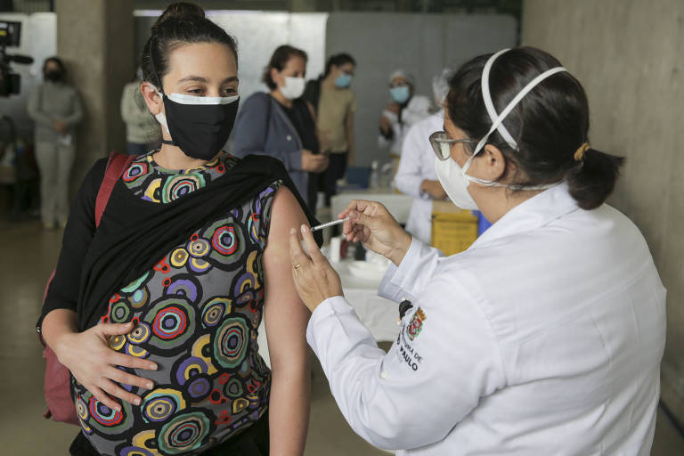 Uma mulher grávida de máscara recebe a vacina pelas mãos de uma profissional da saúde. 