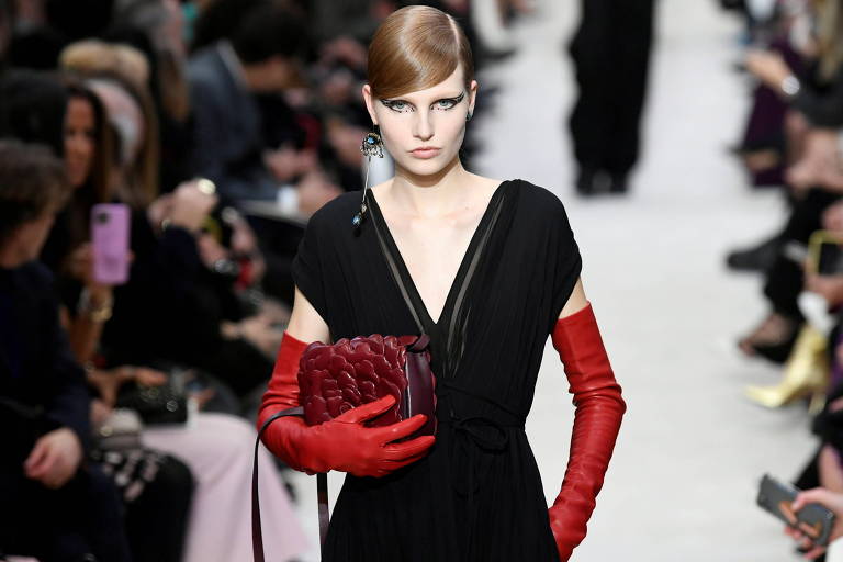 Mulher branca usa vestido preto, luvas vermelhas e bolsa de couro cor de vinho