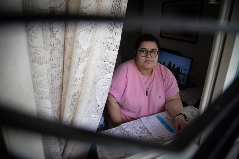 De camiseta rosa, óculos e cabelos escuros presos, Mariana é vista através da janela de sua casa