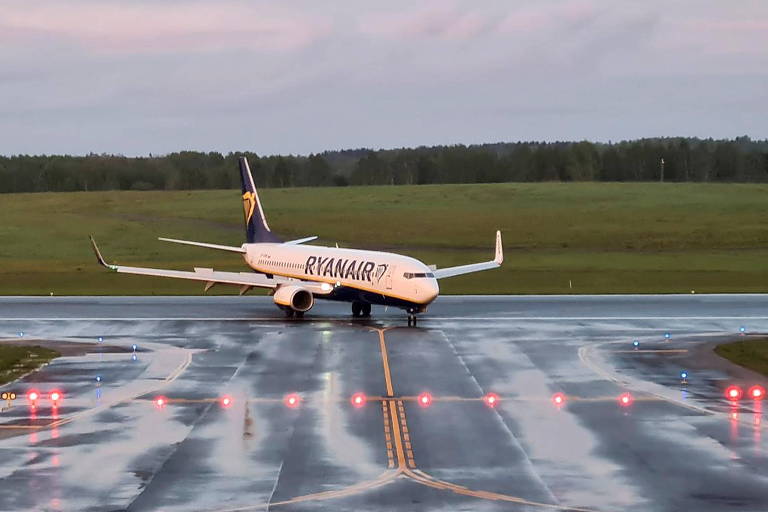 Avião branco da Ryanair vira em pista de pouso molhada pela chuva, com uma fileira de luzes vermelhas à frente, no chão