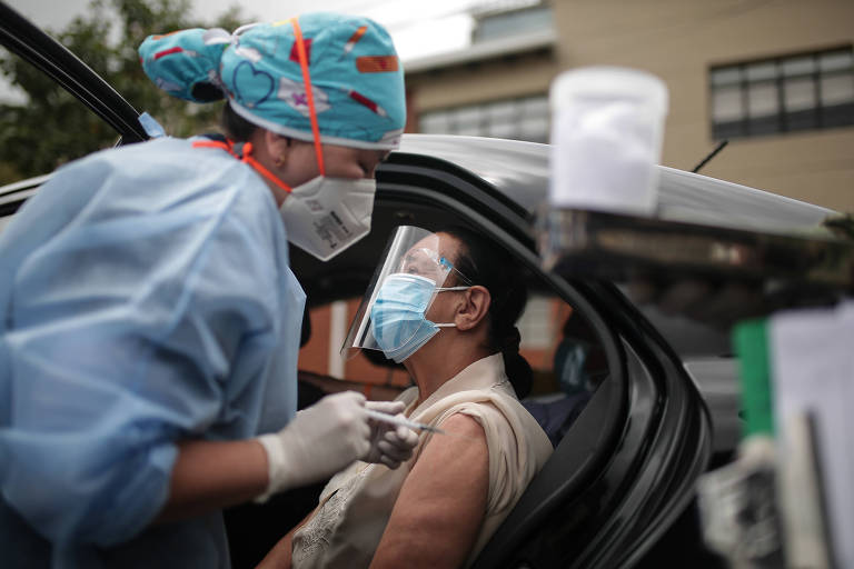 Enfermeira com roupa de proteção aplica injeção em braço de mulher em carro