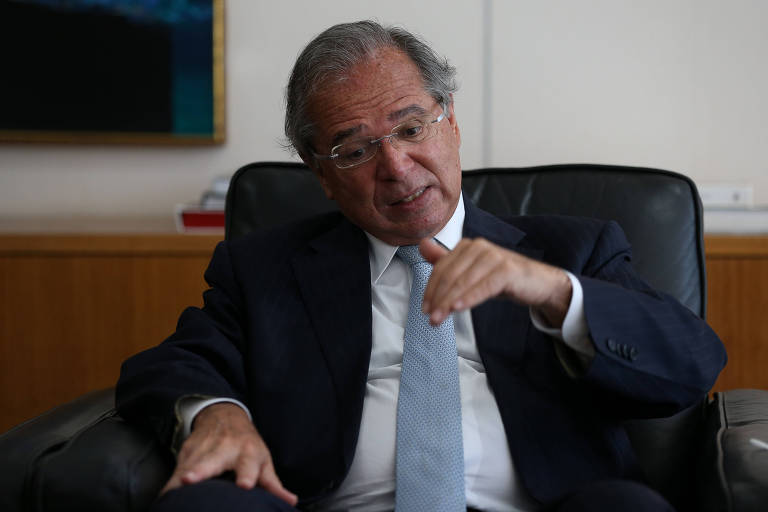 O ministro da Economia, Paulo Guedes, sentado em seu gabinete, gesticula apontando a mão para baixo