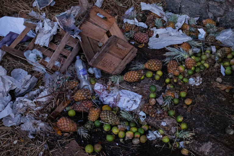 Abacaxis e laranjas misturados a lixo de caixas de madeira, papéis e sacolas jogados no chão