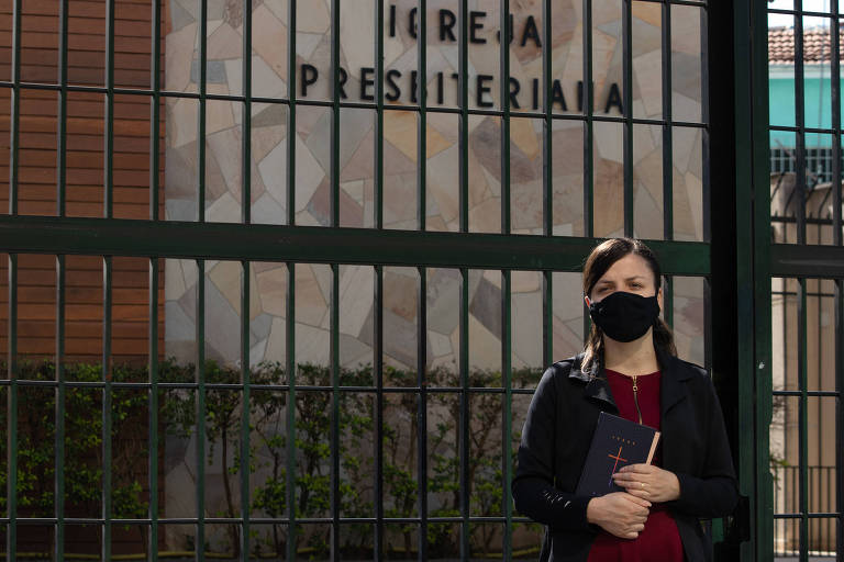 Ela está parada em frente à grade da igreja, de blusa vermelha, casaco e máscara pretos, um livro na mão; ao fundo no muro de pedra se lê IGREJA PRESBITERIANA