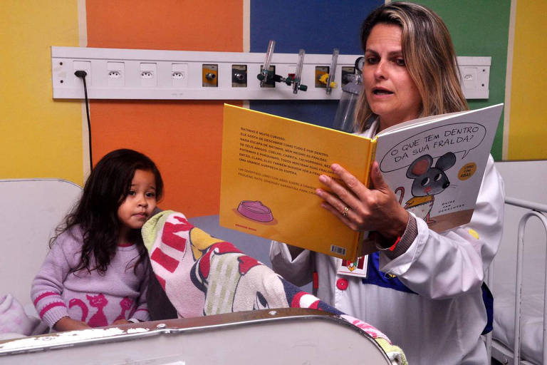 Mulher loira lê livro para menina de roupa rosa e cabelos escuros compridos em cama hospitalar