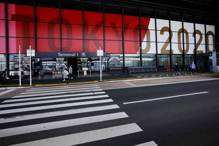 Rua dentro do aeroporto praticamente vazia e iluminada; na parede ao fundo está projetada uma imagem iluminada da bandeira do Japão com as palavras Tokyo 2020