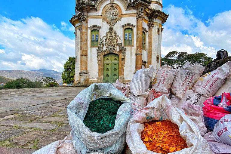 Imagem mostra sacos brancos com serragem colorida (cada saco de uma cor), com igreja ao fundo, em Ouro Preto