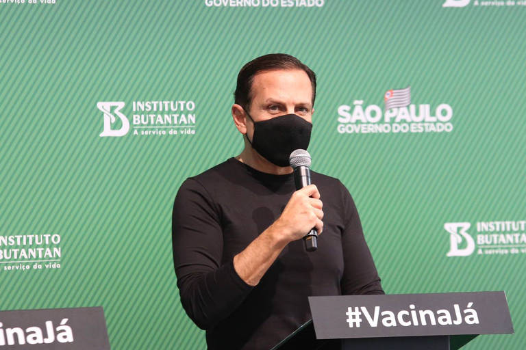De camiseta preta, máscara preta e cabelos escuros curtos, segurando um microfone, João Doria fala em púlpito preto com os dizeres "Vacina Já"