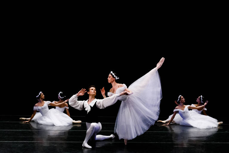 Foto horizontal de bailarinos clássicos, com dançarinas do corpo de baile no chão e casal ao centro