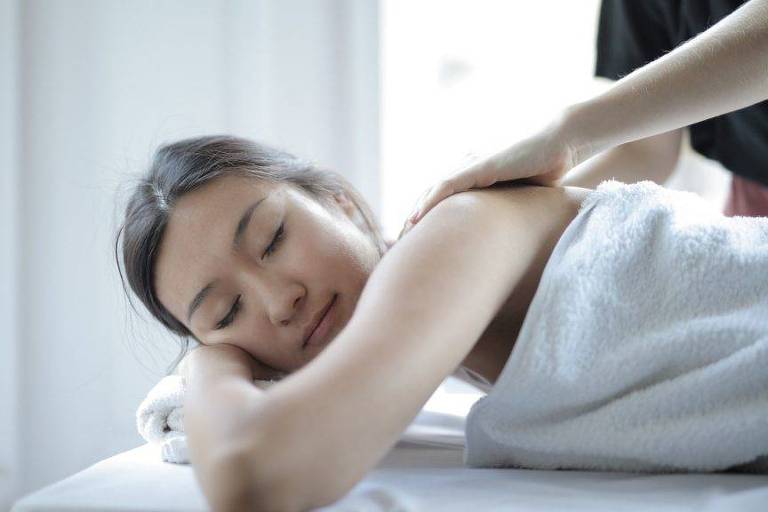 Mulher deitada em mesa de massagem, de olhos fechados e cabelos presos, com mãos em suas costas.