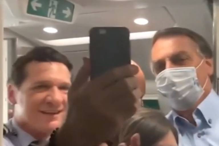 Piloto da Azul segura o celular com a mão direita e para ao lado de Jair Bolsonaro para tirar uma selfie dentro do avião.