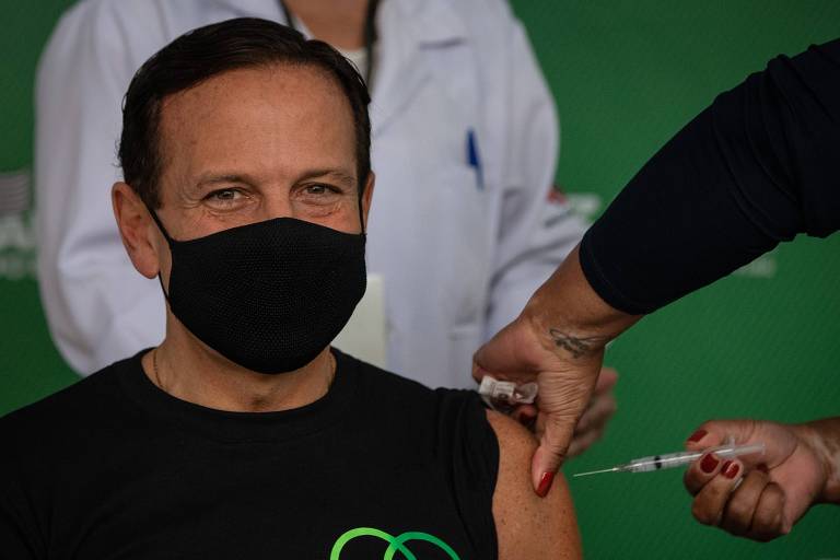 Doria está de máscara e camiseta pretas; na camiseta, lê-se "Vacina pra todos". Ele está recebendo uma injeção