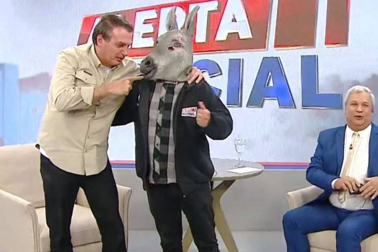 Sem máscara, Bolsonaro está em pé abraçando ao lado um homem vestido com uma cabeça de burro. O apresentador do programa está sentado próximo a eles