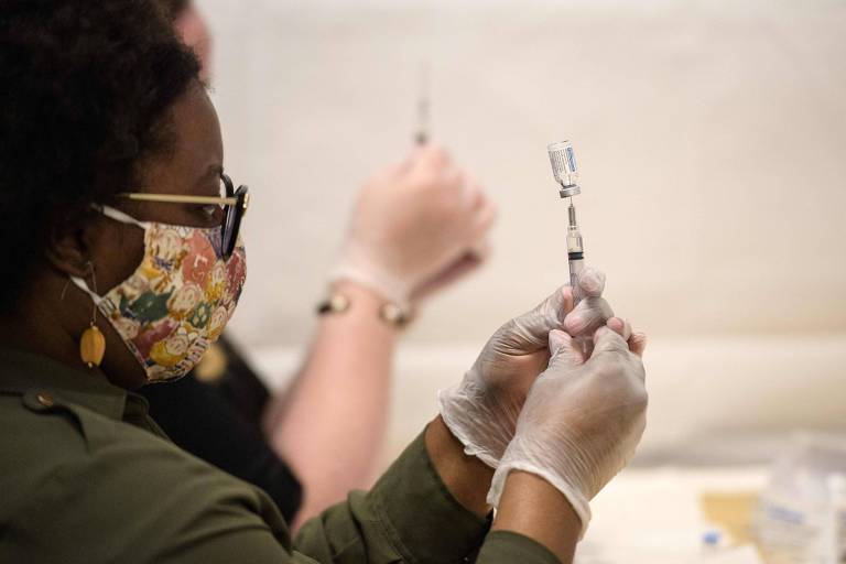 Imagem em primeiro plano mostra enfermeira de máscara segurando uma seringa espetada em uma ampola de vacina