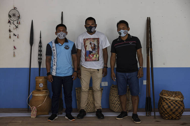 Três homens em pé encostados contra a parede com ferramentas indígenas ao fundo