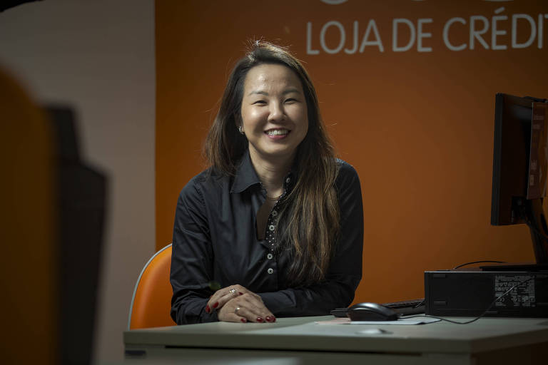 Milene está sentada em uma mesa de escritório e sorri para a foto à frente de uma parede laranja