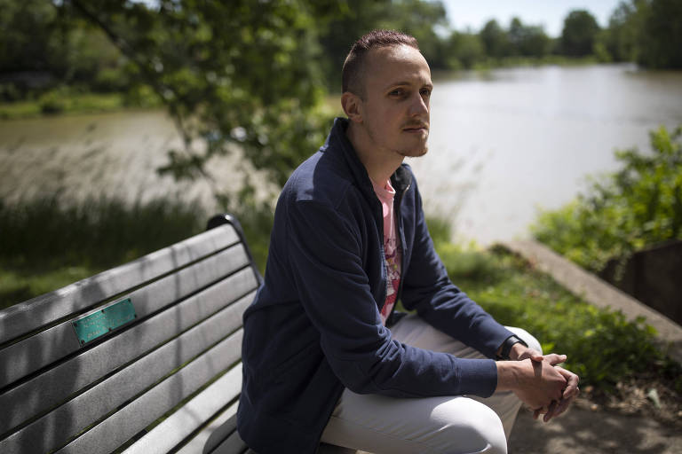 Homem branco, de bigode e cavanhaque, está sentado em um banco de madeira à margem de um lago