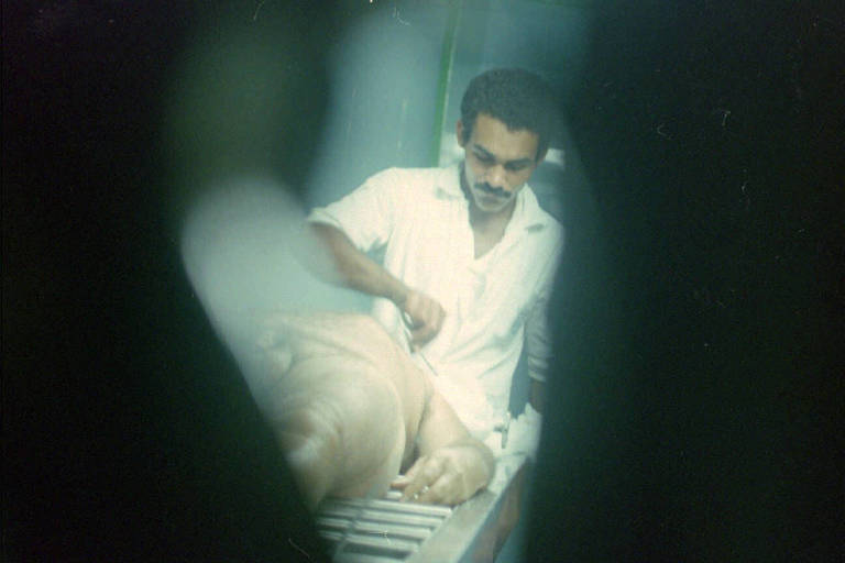 Visto por uma fresta, corpo do empresário é examinado por funcionário no IML (Instituto Médico Legal) de Maceió