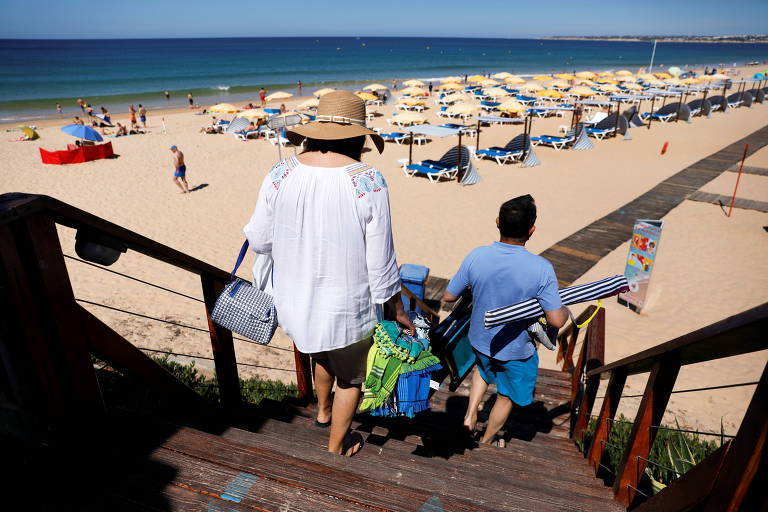 Homem, criança e mulher de chapéu descem escada em direção a praia de faixa larga de areia, com fileiras de guardassóis brancos