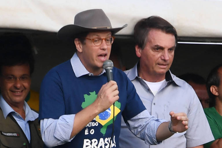 Salles, com chapéu de cowboy e camiseta que tem um mapa e estampa da bandeira do Brasil, ao lado de Bolsonaro, que parece olhar para baixo e para frente