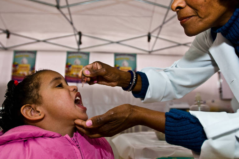 Criança é fotografada de boca aberta, enquanto uma enfermeira pinga a vacina de poliomielite.