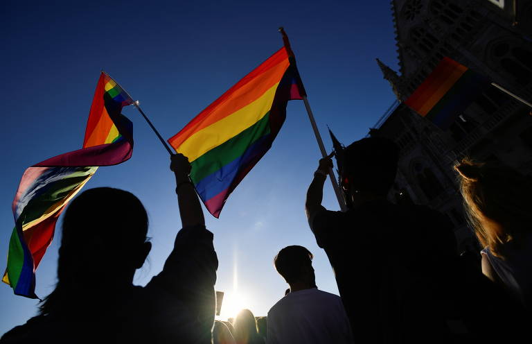 fotografia colorida mostra pessoas empunhando a bandeira do arco-íris durante manifestação 