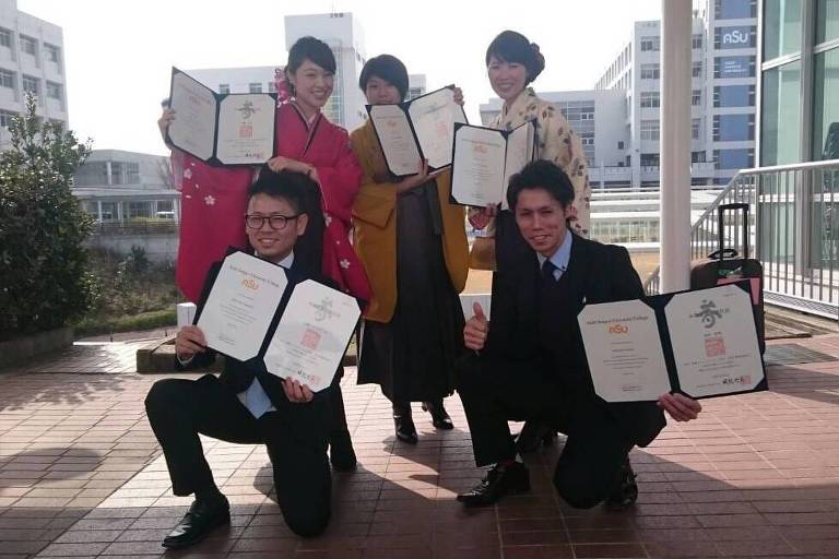 Dois homens de terno (agachados) e três mulheres (em pé) com roupas típicas japonesas mostram seus diplomas